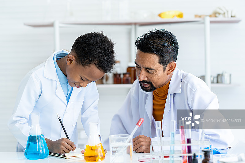 在化学教室里有许多实验工具。一个年轻的非洲男孩和穿着白大褂的男老师互相帮助做实验。一个男孩记录数据，老师看着他写的课本。图片素材