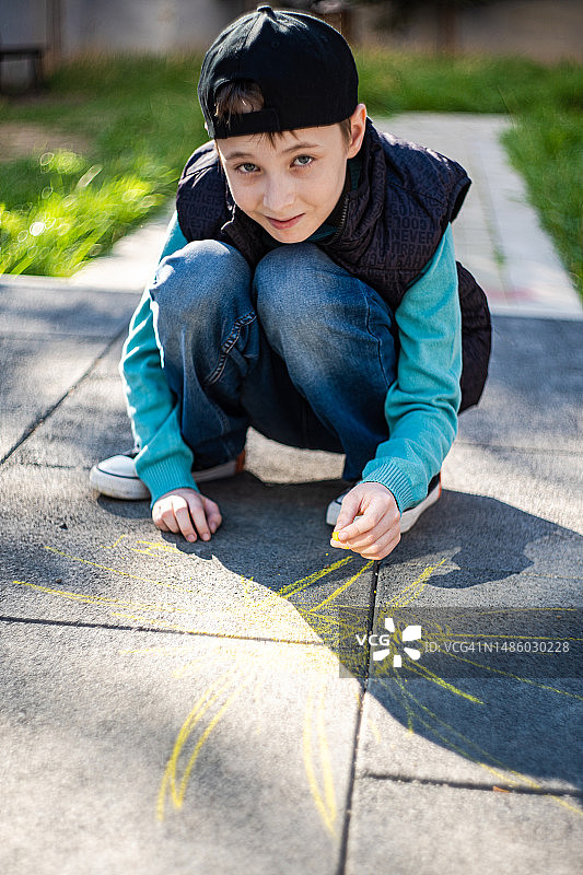 一个微笑的男孩蹲在人行道上用粉笔画画图片素材