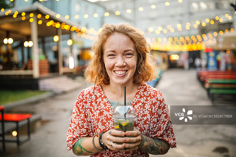 拥抱真实:笑着逗人的红发女人在街上享受着吸管味的柠檬水。图片素材