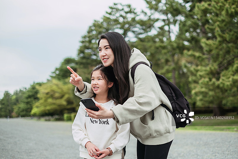 年轻的亚洲母亲和她的女儿在京都的公园里。以树木为背景，向远处眺望并指向远方。旅游概念，母女时刻，家庭，假期，假期，家庭周末活动，规划和成长。图片素材