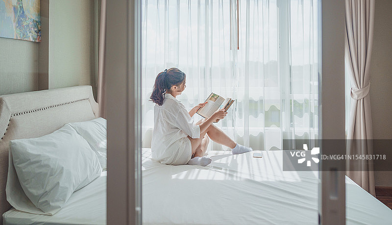 放松年轻的亚洲女人享受她的咖啡，而坐在床上。女人在家里的卧室里边看书边喝咖啡图片素材