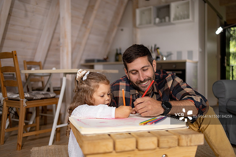快乐的父亲和女儿在家里放松时在笔记本上画画。图片素材