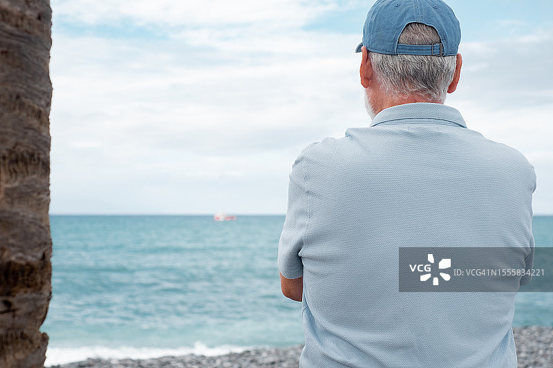 身穿休闲polo衫的老人站在沙滩上望着大海的地平线，戴着帽子的退休老人享受着闲暇时光和退休生活图片素材