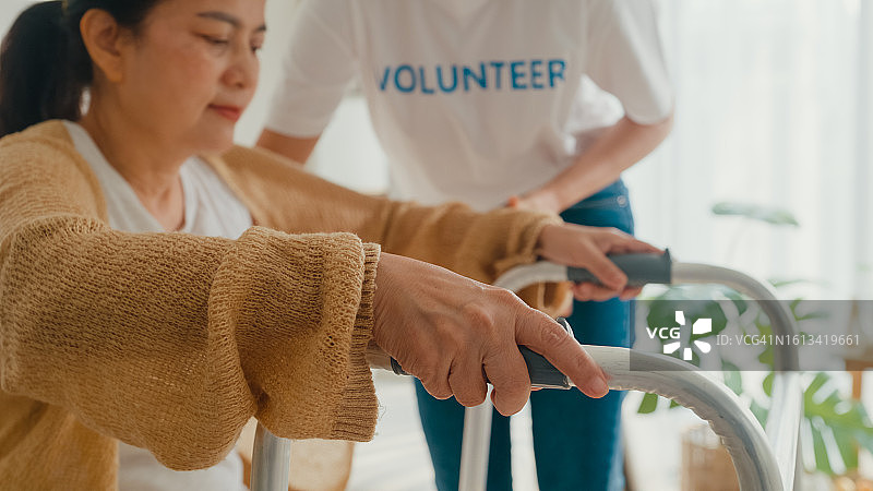 年轻的亚洲女性志愿者在家中教老妇人走路用助行器，保重手扶住客厅中的老年病人。志愿医疗保健。图片素材