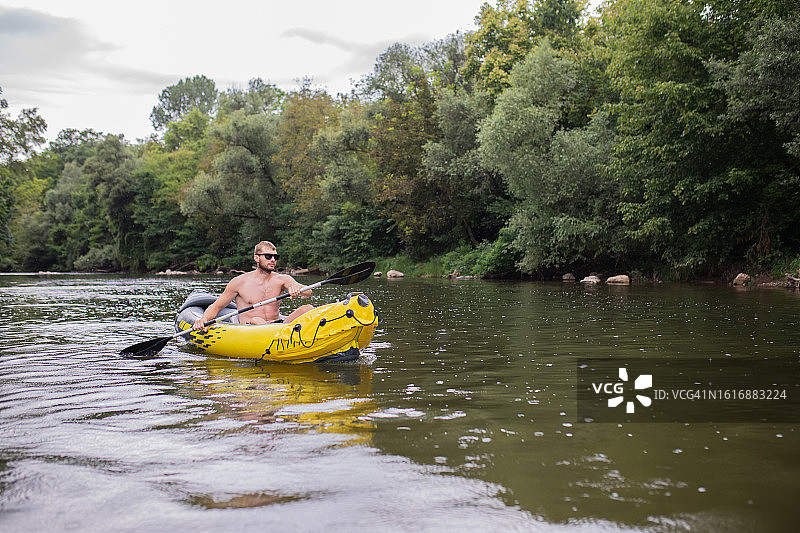 年轻的爱冒险的大胡子男子正用桨划着他的黄色充气独木舟在河上划船。图片素材