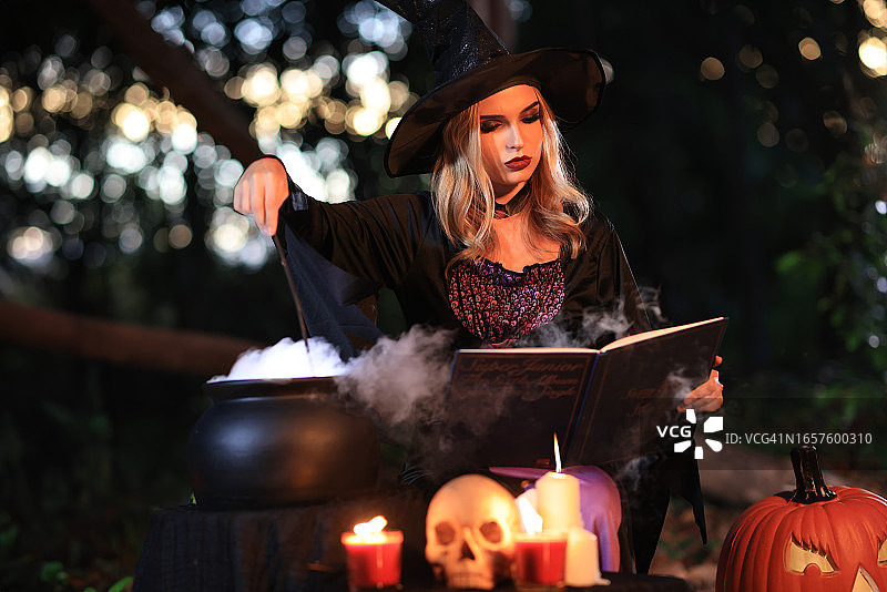 幻想中的女女巫会变魔术，手里拿着一根魔杖和一本魔法书，念着从沸腾的大桶里冒出来的白魔法烟。万圣节的主题。女巫在万圣节那天举行仪式图片素材