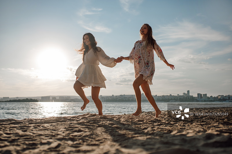 两位穿着迷人背心裙的女士手牵着手沿着沙滩奔跑图片素材