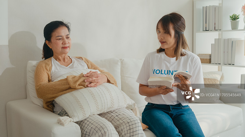 年轻的亚洲女性志愿者在家里客厅的沙发上和快乐的老妇人坐在一起读书。志愿医疗保健。图片素材