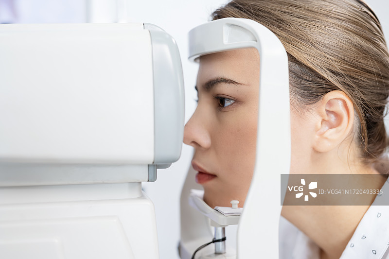 照顾你的眼睛健康，减少眼部疾病，进行全面的眼科检查。一位耐心的年轻女子在眼科诊所里把下巴靠在光学仪器上检查眼睛。定期进行眼科检查。图片素材