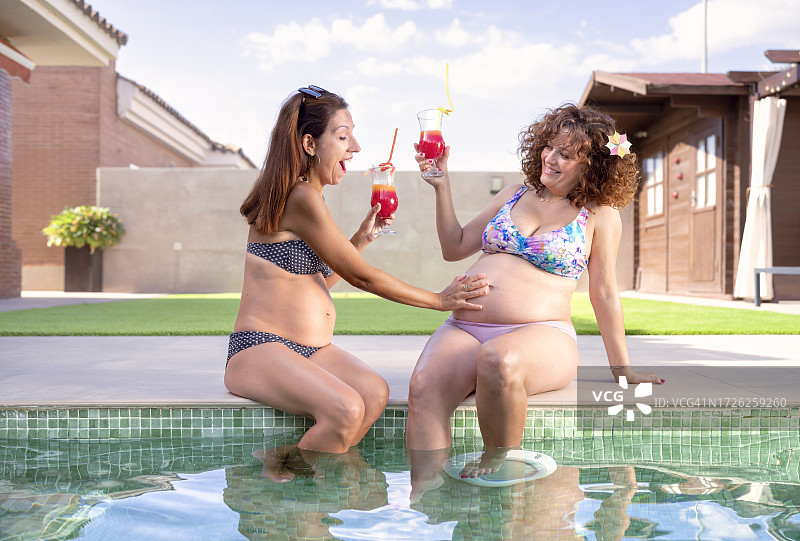 两个孕妇朋友在自家花园的游泳池里举杯庆祝自己怀孕了，其中一个抚摸着另一个的肚子图片素材