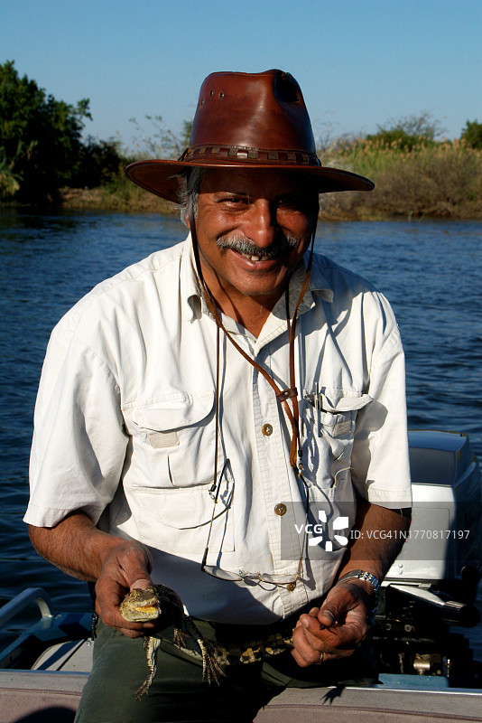 博茨瓦纳，奥卡万戈河上，一名微笑的男子抱着一只小鳄鱼坐在船上图片素材
