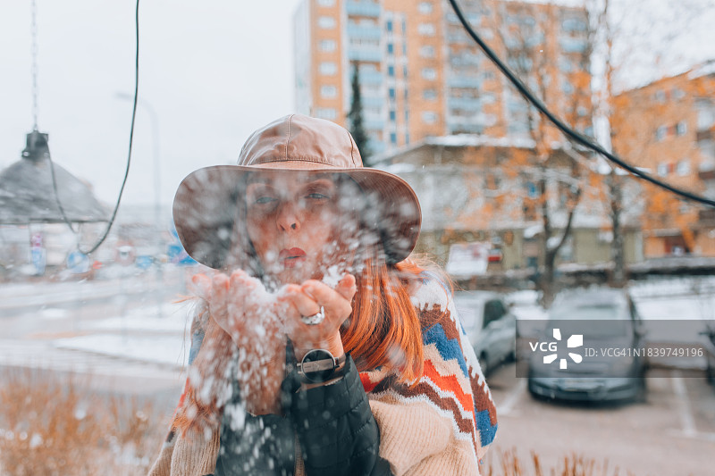 迷人的女人穿着彩色雨披，戴着大沿帽，吹着手中的雪朝镜头走去图片素材