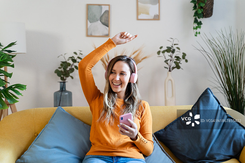 快乐的年轻女子坐在家里的沙发上，戴着耳机听音乐图片素材
