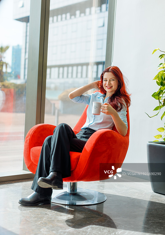 办公室休息的幸福:年轻女商人的肖像休息，舒适地坐在红椅子上，喝着咖啡，放松。在现代明亮的办公室休息时，她沐浴在阳光中，享受着窗外的阳光图片素材