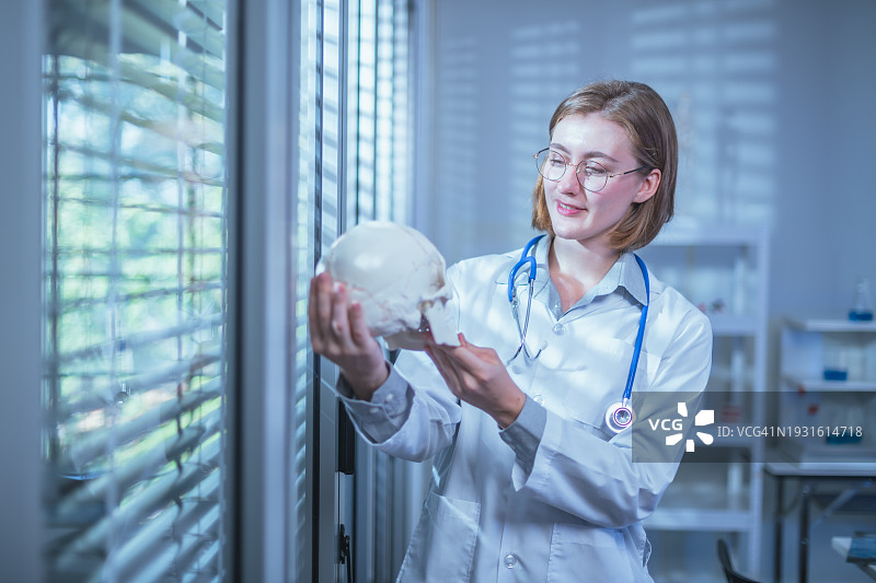 这位年轻的女医生站在头骨模型旁边，她的表情沉思而专注，因为她检查了人类头骨的复杂细节，可能是在精确而谨慎地考虑诊断或治疗计划。图片素材