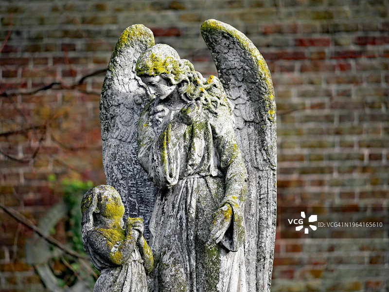 英国伦敦布朗普顿公墓的古代雕像，描绘了一个天使和一个孩子。没有人。背景:风化的砖墙。阳光。自然的颜色。图片素材