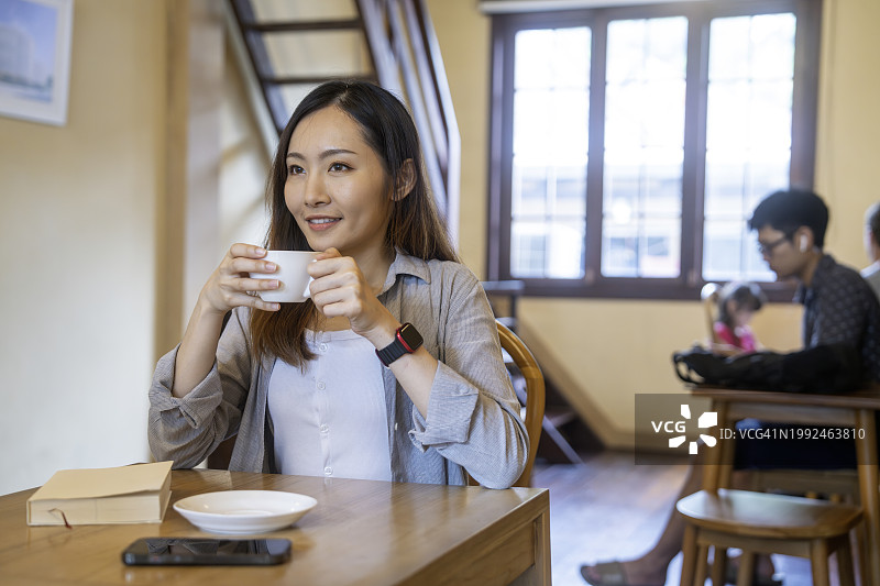 愉快的亚洲年轻女子喝热咖啡或茶，坐在咖啡馆享受它图片素材