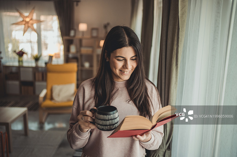 年轻漂亮的女人在家里看书，喝着一杯热茶图片素材