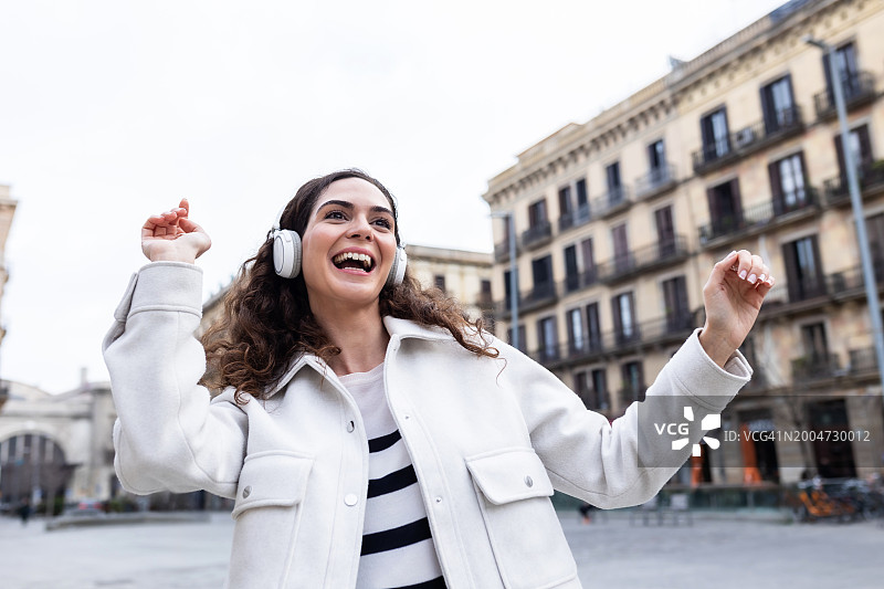 快乐的年轻成年女子一边戴着无线耳机享受网络音乐，一边在城市街道上跳舞。图片素材