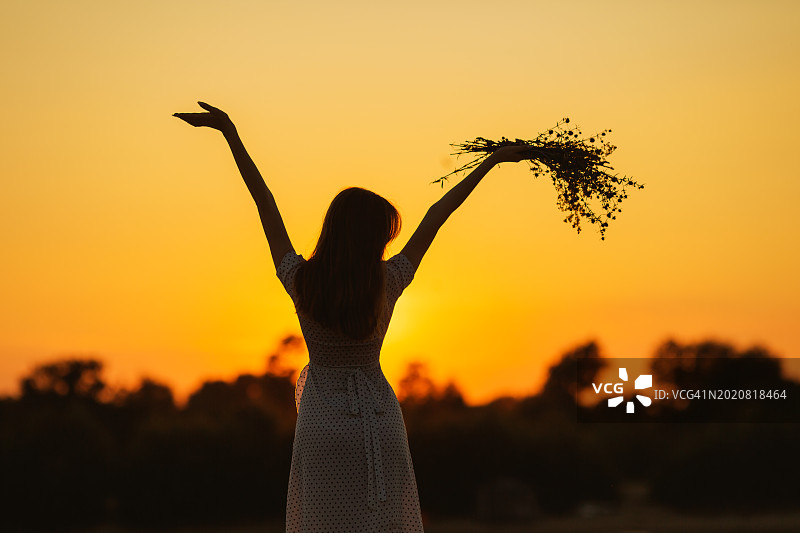 金发女人穿着裙子，手捧一束野花。夕阳背景下的剪影。图片素材