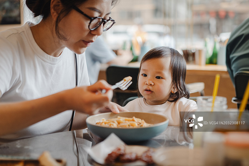 在一个惬意的下午，年轻的亚洲母亲在餐厅里和她蹒跚学步的女儿分享意大利面。全家外出就餐的生活方式。亲情之爱，关怀与纽带的理念图片素材