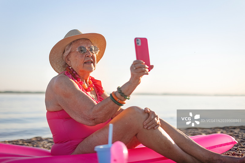 一位老妇人手持智能手机，坐在粉红色的泳池浮子上，在沙滩上自拍。一位老年妇女在海边享受暑假。退休后旅行的概念。图片素材
