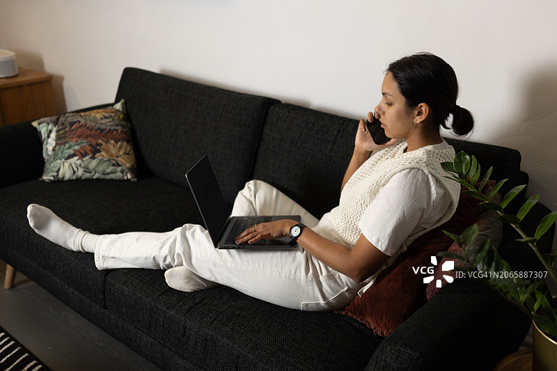 坐在沙发上使用笔记本电脑和智能手机在家工作的女性图片素材