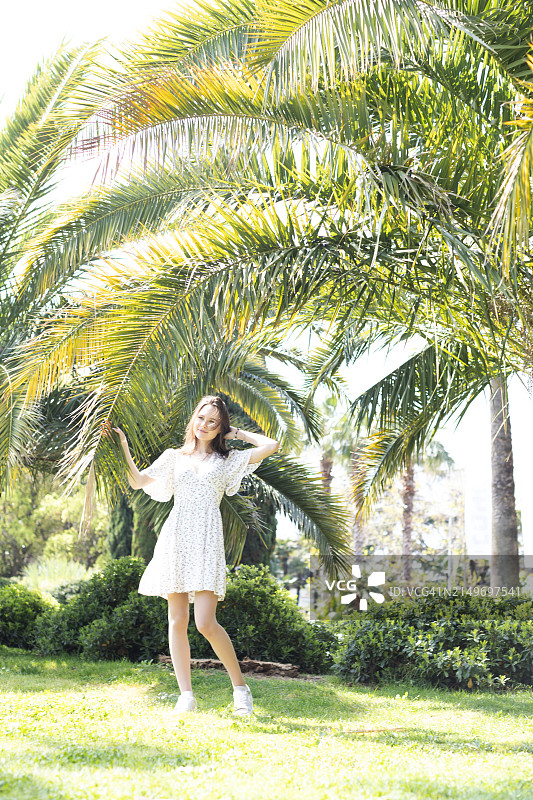 美丽的年轻女子穿着浅色的夏装，在棕榈树的背景下散步图片素材