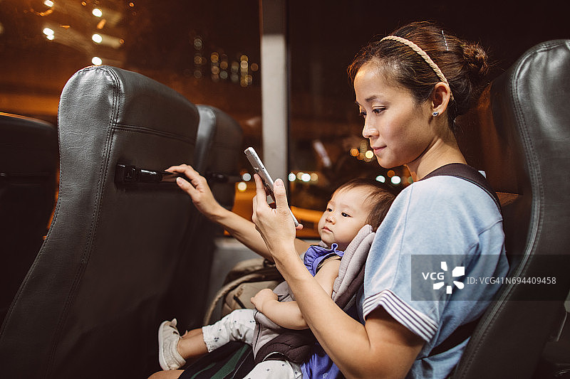 年轻漂亮的妈妈在公交车上用智能手机抱着她的孩子。图片素材