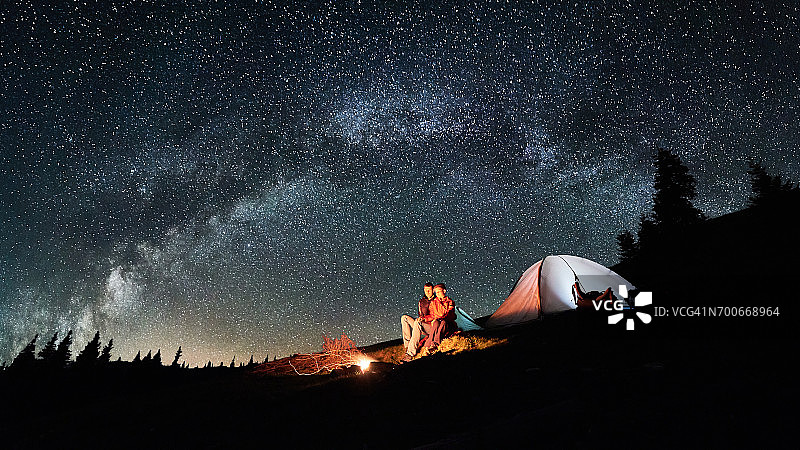 晚上露营。浪漫的游客情侣们在明亮的帐篷旁的篝火旁休息，夜空中满是星星和银河。天体摄影术。图片宽高比16:9图片素材