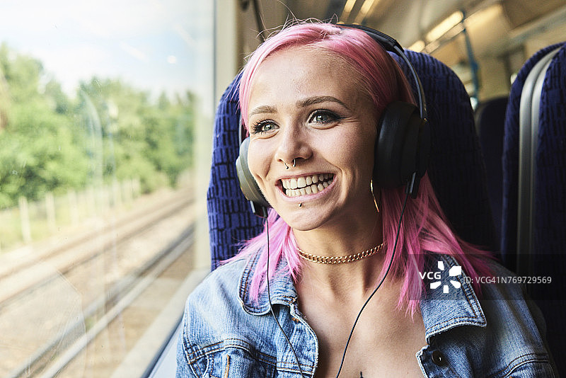 快乐的年轻女子与粉红色的头发听音乐，同时乘火车旅行图片素材