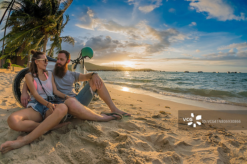 一对夫妇——一个留着胡子的男人和他英俊的女朋友——倚在沙滩上的摩托车上，看着海边的日落图片素材