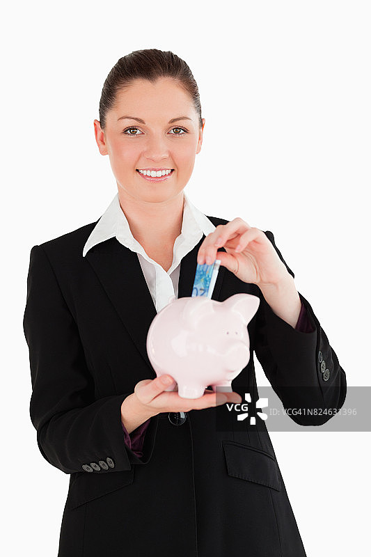 一个迷人的穿着西装的女人把一张钞票放进一个粉红色的储蓄罐图片素材