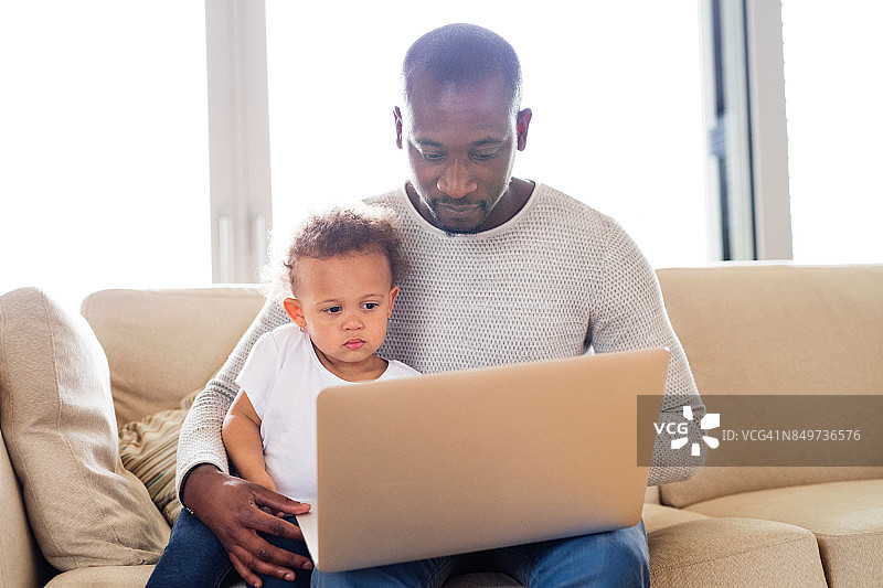 非裔美国父亲和小女儿在家拿着笔记本电脑。图片素材