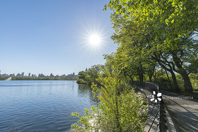 春天的阳光照亮了水库、新鲜的绿树和纽约中央公园水库边的斯蒂芬妮和弗雷德舒曼跑步道。中央公园西历史区可以看到后面的水库。图片素材