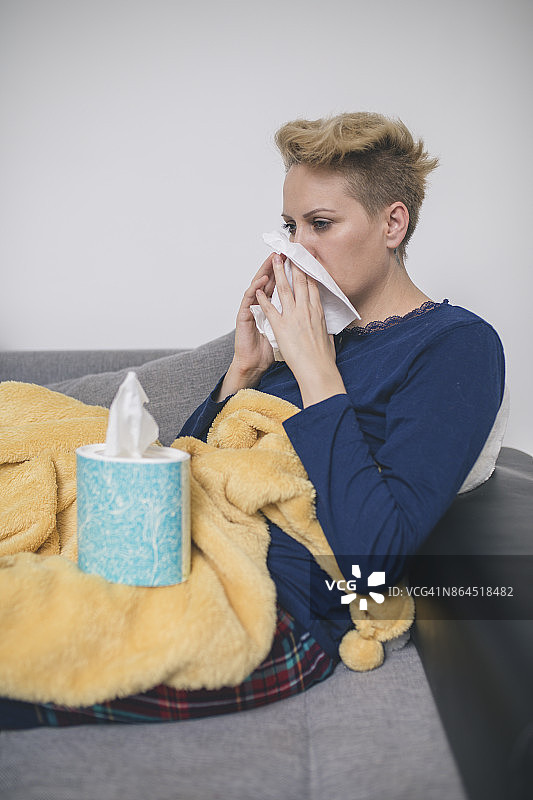 一个生病的年轻女子因感冒坐在家里的沙发上，她盖着一条毯子，擤着鼻子图片素材