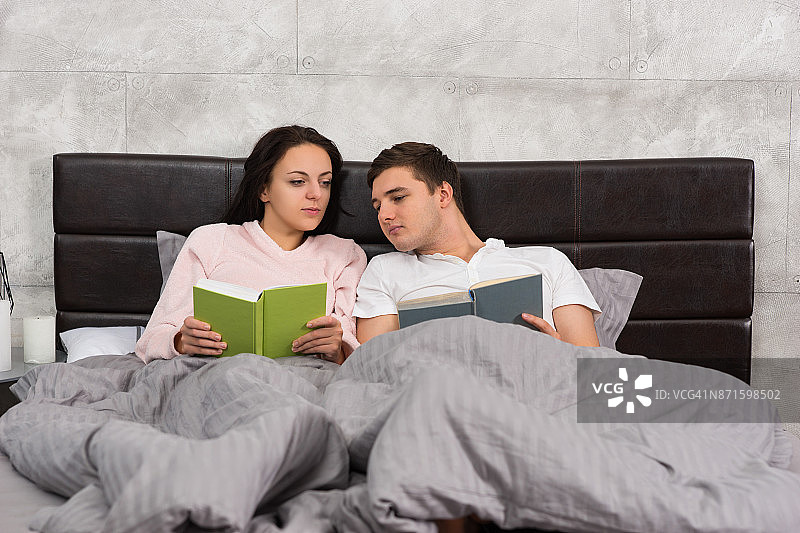一对年轻夫妇穿着睡衣躺在床上读对方的书图片素材