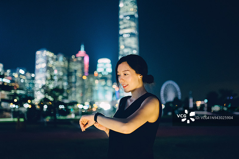 年轻的亚洲女性在智能手表上查看时间，背景是明亮的城市夜晚图片素材