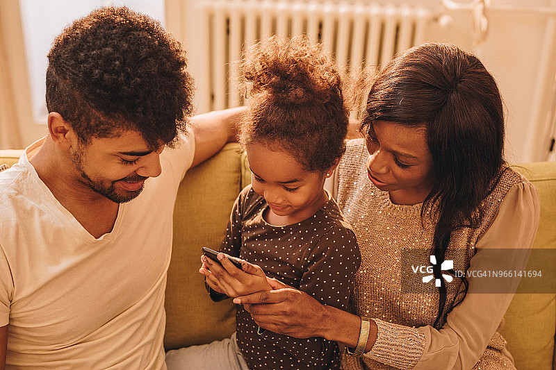微笑的黑人女孩和她的父母玩得开心，而使用智能手机。图片素材