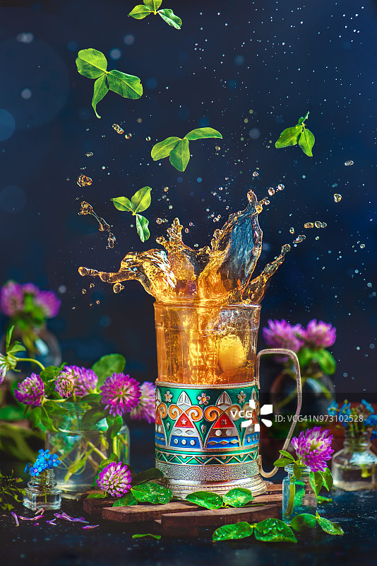 茶叶飞溅在搪瓷杯架与飞落的叶子和水滴。有三叶草茶和花的静物画。黑暗图片素材