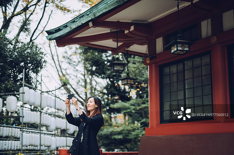 快乐的亚洲女性游客在镇上观光时，用手机在传统的日本房屋前自拍图片素材