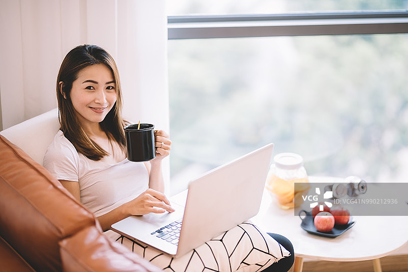 一位亚洲华人女性坐在客厅的沙发上看着笔记本电脑，她的移动设备拿着一杯咖啡图片素材