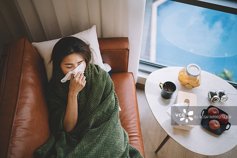 一名生病的华裔女性躺在客厅的沙发上，身上盖着毯子，用纸巾擦着鼻子图片素材