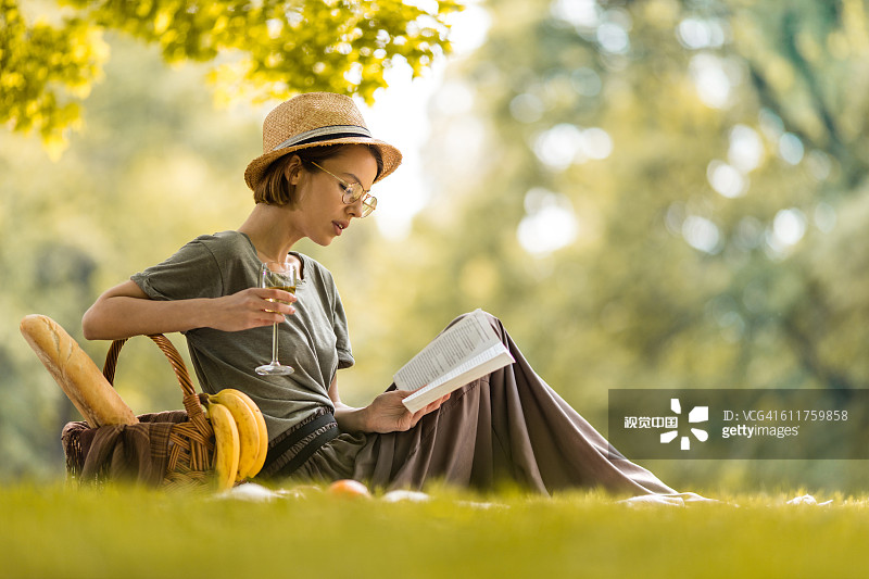 年轻女子放松与葡萄酒杯和阅读书籍在野餐。图片素材