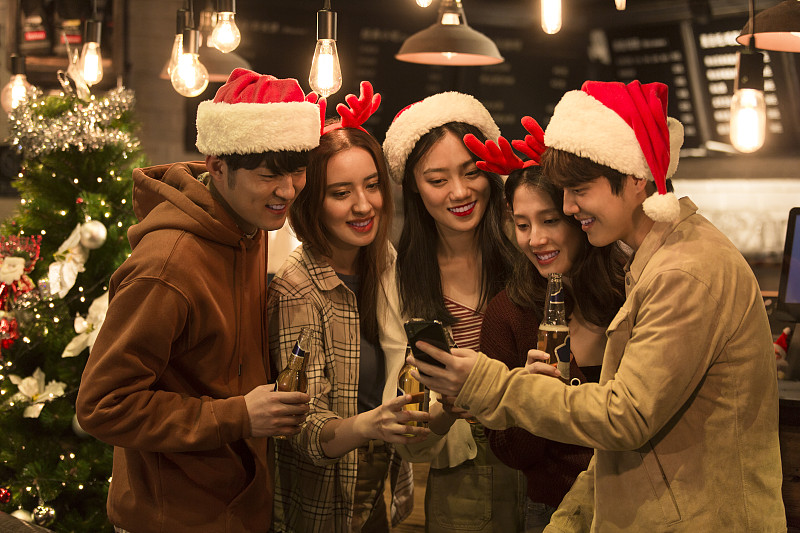 五个青年男女戴着圣诞帽和发卡边喝啤酒边玩手机在酒吧庆祝圣诞节图片下载