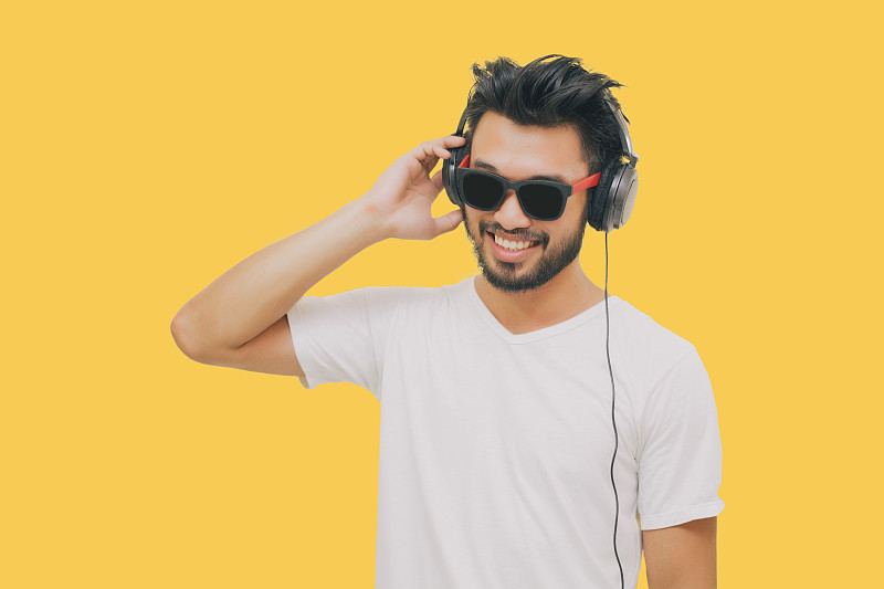 亚洲英俊的男人，有胡子，微笑和大笑，用智能手机听音乐的黄色背景耳机图片下载