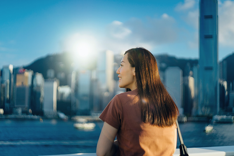 在一个阳光明媚的日子里，一名年轻亚洲女子在维多利亚港海滨漫步，微笑着欣赏香港城市的天际线图片下载