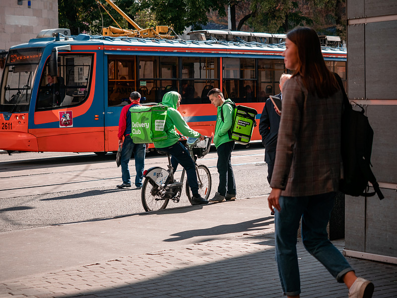 两名身穿制服，背着印有绿色快递俱乐部品牌的背包的快递员在一个电车站旁边交流。带有标志和标志的保温袋，送食品的公司图片下载