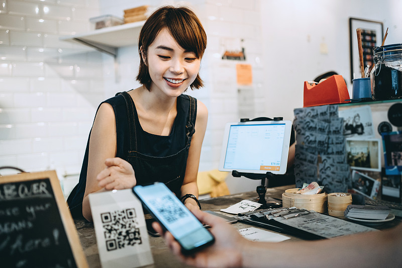 顾客扫描二维码，用智能手机进行快速、方便的非接触式支付，前面是微笑的咖啡师图片素材