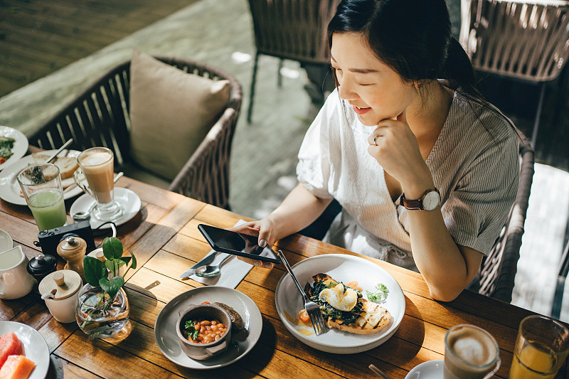 微笑的年轻亚洲妇女使用手机在一个户外餐厅吃早午餐对美丽的阳光图片下载
